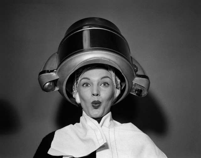 1950s woman under hair dryer with towel on shoulders and hair net looking at camera